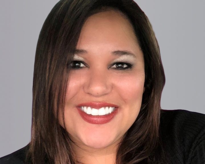 Portrait of a smiling woman with long brown hair, wearing a black jacket, against a gray background.