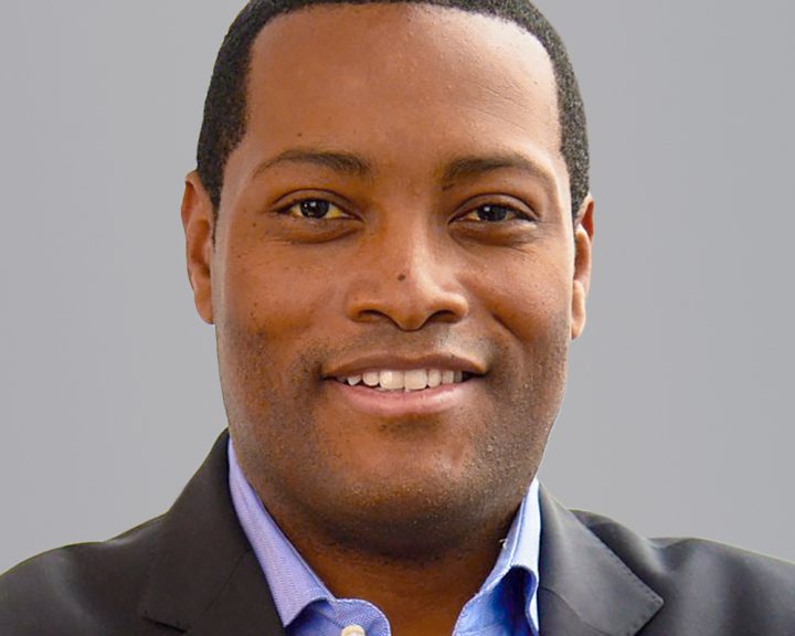 Portrait of a smiling man wearing a blue shirt and black blazer against a grey background.