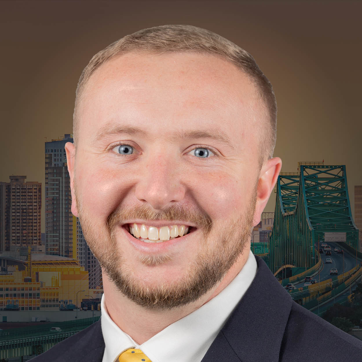 Man smiling in a suit and tie, with a city skyline and green bridge in the background.